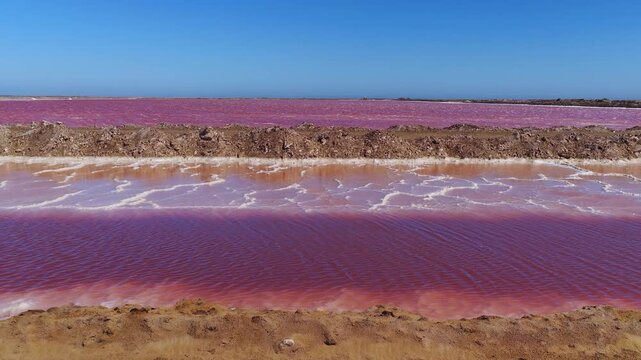 Aerial view of pink salt evaporation ponds near Walvis Bay, Namibia. Camera glides over basins, ripples, mud banks, salt crusts, and earthen berms at midday.