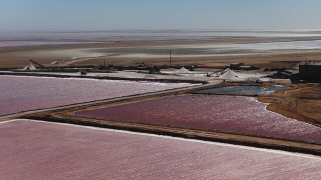 Aerial view of Namibia salt pans near Walvis Bay shows pink and rose ponds, white salt piles, conveyor belts, and buildings in midday sun, with shimmering motion.
