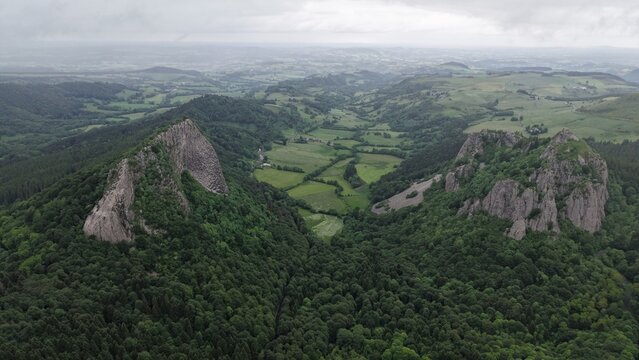 Aerial view of the Roches Tuiliere et Sanadoire, two volcanic rock formations rising above a lush green valley with dense forests in Puy-de-Dome, Auvergne-Rhone-Alpes, France.
