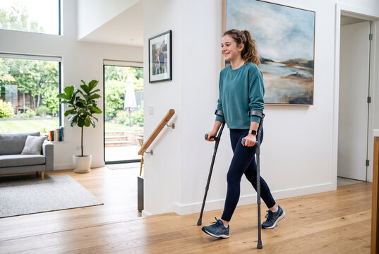 A young woman with crutches walks in a bright room of a modern house. She has a smile on her face and is wearing casual clothes. The room has a large window and plants