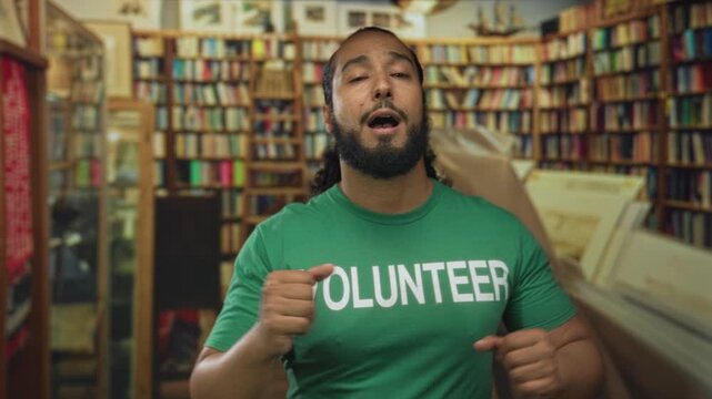 Man in volunteer shirt clenches fist near bookshelves in building aisle; community service determination.