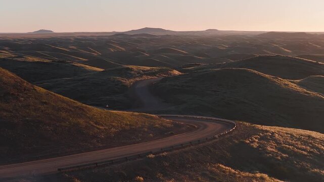 Aerial view shows low grass dunes in warm golden hour light, a curving dirt road with metal guardrail, tiny figures at a bend, and rocky knobs near the Skeleton Coast.