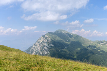 Fototapeta premium In Italy, a green mountain ridge with a pale rocky face rises across Monte Baldo, surrounded by meadow, forest patches and scattered summer clouds.