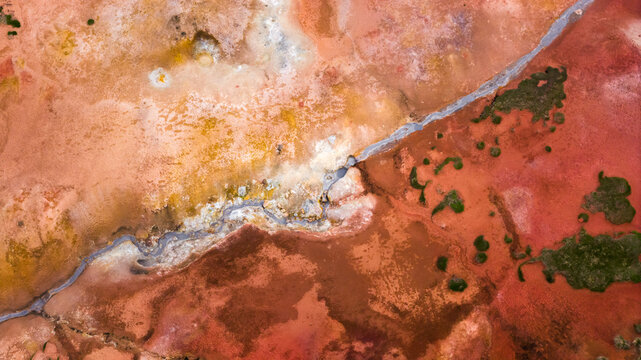 Aerial view of geothermal terrain featuring vibrant red and orange mineral deposits, steaming water channels, and patches of green moss in Husavik, Nordurthing, Iceland.