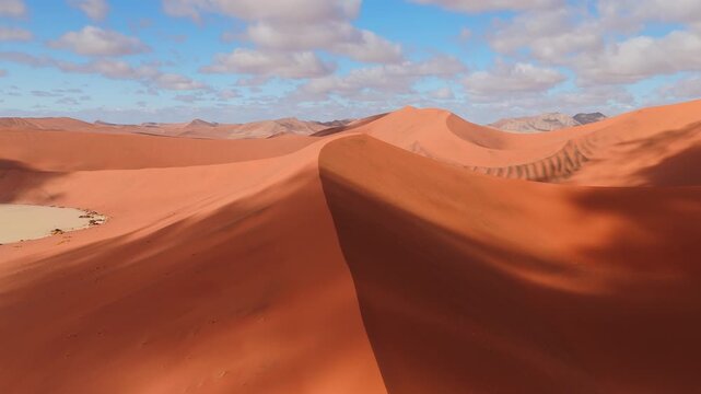 Aerial camera sweeps along a sharp dune ridge in Sossusvlei, Namibia, showing ripple patterns, a pale clay pan, and rocky ridges under bright sky and warm sand.
