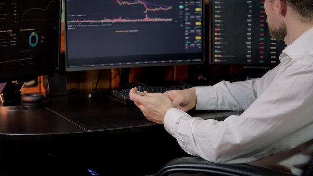 A man sits at three monitors, analyzing market data and counting cash in a dimly lit office