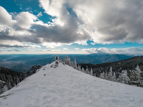 Ground level photograph of a female hiker with backpack standing on a snowy mountain peak looking over the winter valley landscape in Cozia Massif, Romania.