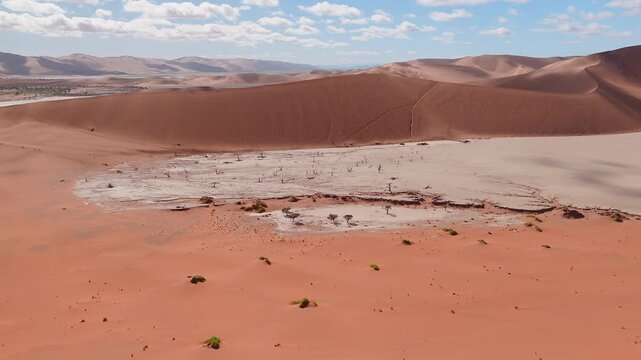 Aerial view of Deadvlei in Sossusvlei, Namibia shows a white clay pan, dead camel thorn trees, rust red dunes, tiny visitors on ridges, and a smooth forward sweep.