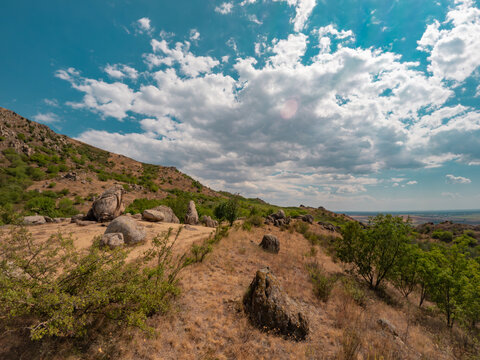 Wide angle shot of the granite monoliths known as Dinosaur Eggs rock formations overlooking arid hills under a cloudy blue sky in Macin Mountains, Dobrogea, Romania.