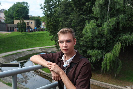 A young blond guy in brown clothes stands on a city bridge over river.