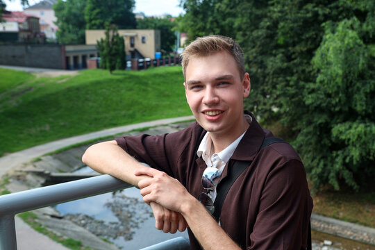 A young blond guy in brown clothes stands city bridge over a river.