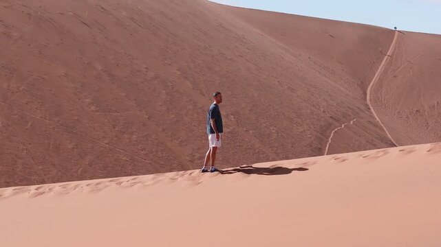 Aerial shows a lone visitor on a red dune crest above Deadvlei in Sossusvlei, Namib Desert, Namibia. Bright daylight, crisp shadows, wide frame, groups below.