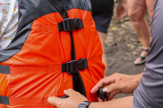 Orange life jacket being fastened by hands on outdoor riverbank with people in background, excited safety preparation for water activity