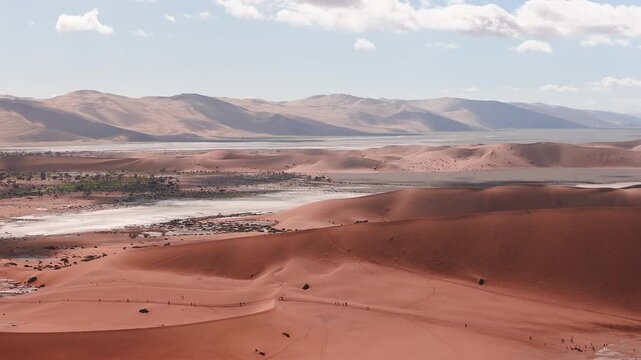 Aerial view shows rust red dunes, pale salt pans, Sossusvlei in Namib Naukluft Park, distant ranges, scattered vegetation, tiny people on ridges, soft clouds, daytime sweep.