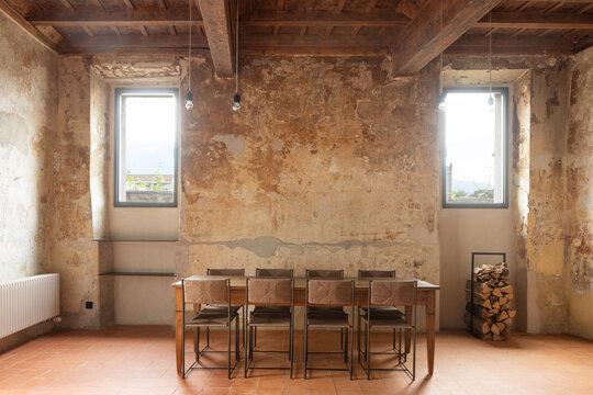 The interior of a beautiful dining room seating eight people. A large table with eight chairs, and old, peeling walls. Daylight streams in through two windows.