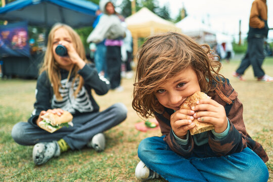 Two children sitting on grass eating sandwiches at an outdoor festival