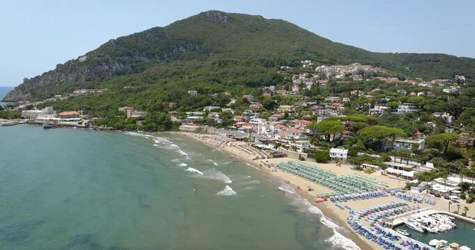Aerial view of the beach at San Felice Circeo in Italy, featuring organized sun loungers on golden sand at the foot of Mount Circeo's green slopes by clear sea waters on a sunny summer day.
