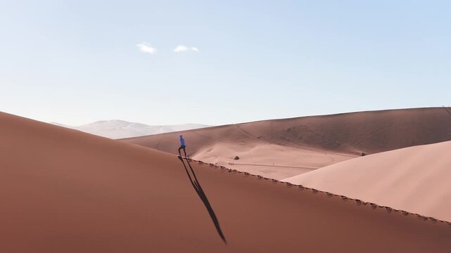Aerial view shows a lone walker on orange red dunes in Sossusvlei, Namib Desert, Namibia. Footprints cut diagonally as long shadow falls in low golden light.