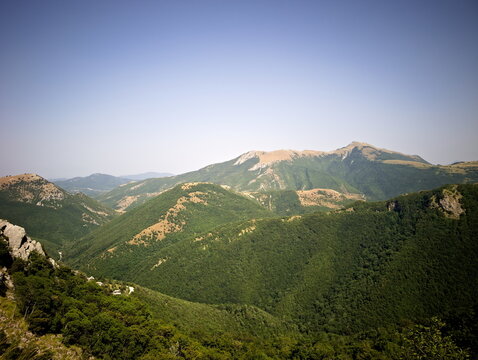 Paesaggio montano con montagne rocciose, foresta verde sui pendii e catena di rilievi che si estende in lontananza sotto un cielo azzurro limpido