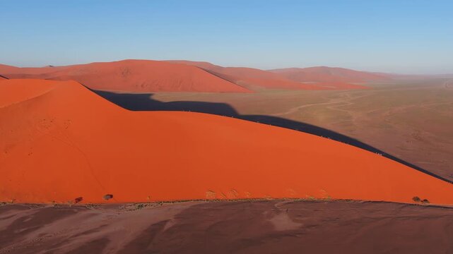 Aerial view of Sossusvlei dunes in Namibia shows a crescent dune 45 casting a deep shadow on the pan as tiny people walk the crest and the camera sweeps in warm low light.