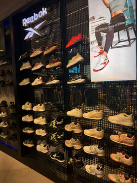 A display of Reebok sneakers showcased on a black mesh wall. Various shoe styles in different colors like red, white, and beige are arranged neatly. The modern retail interior reflects sports fashion.