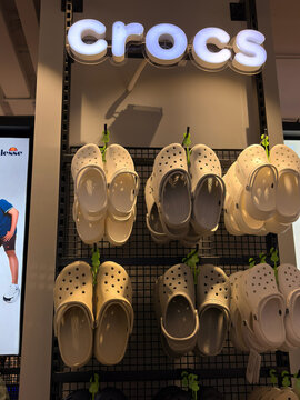 A display of white Crocs footwear arranged neatly on a store shelf. The sign overhead indicates the brand, illuminated in bright letters. The scene has a clean and modern retail environment.