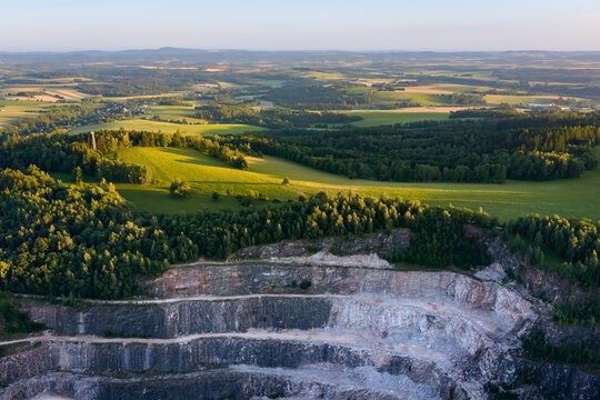 Aerial view of a large open pit stone quarry mine with terraced rock layers surrounded by green landscape during the sunset