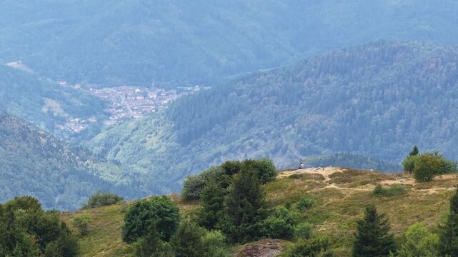 Aerial view of Grand-Ballon mountain with lush green forests and a person sitting on the hill overlooking a distant village in Soultz-Haut-Rhin, Grand Est, France.