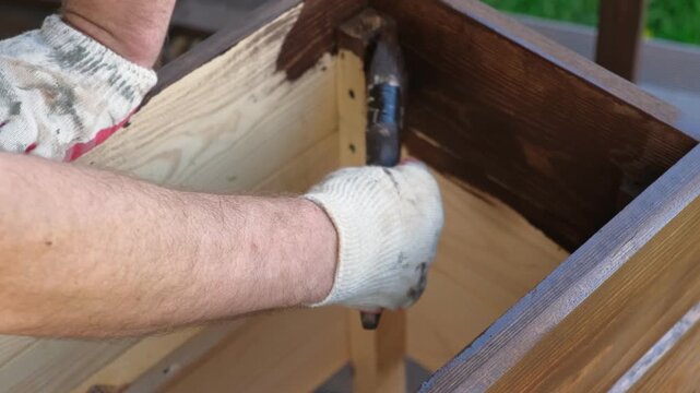 Hand in glove painting wooden box with brown oil stain brush. Worker applying protective varnish on pine timber planks surface, DIY woodwork. Wood staining process. Backyard Woodworking, Maintenance.