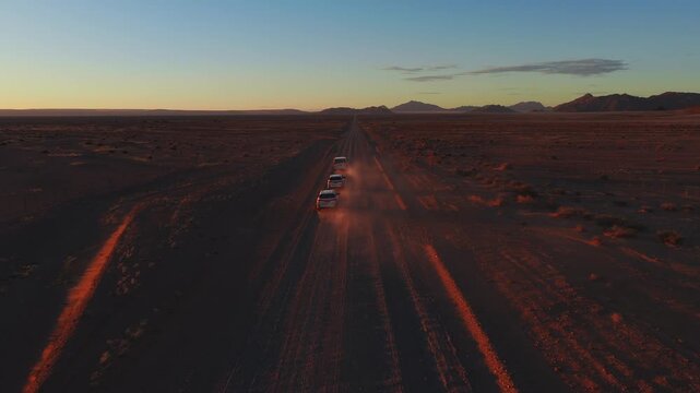 Aerial view shows white 4x4s and a minibus driving on a straight dirt road in the Namib Desert, Namibia, kicking dust toward distant mountains at golden hour.