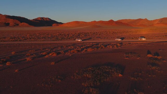 Aerial view shows white SUVs and a van driving a straight gravel road in the Namib Desert, Namibia. Golden hour light casts long shadows over scrub and rocky hills.