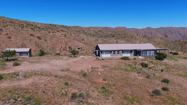 Aerial drone sweep shows a single story concrete house and outbuilding on a dry shrub dotted plateau in Namibia, with a winding dirt track under clear midday sky.