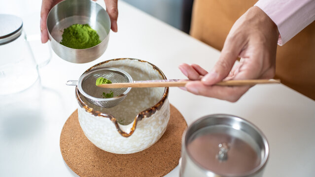 Close-up of hands using a wooden scoop to sift organic matcha green tea powder through a metal strainer into a ceramic bowl. Japanese tea ceremony and cafe culture concept.