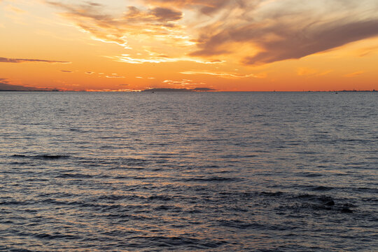 Warm golden sunlight dips below the distant horizon, casting orange reflections across calm ocean waves and a dramatic cloud-filled sky, evoking peaceful, contemplative evening mood by the sea