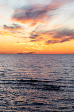 Warm golden sunlight dips below the distant horizon, casting orange reflections across calm sea waves and a dramatic cloud-filled sky, evoking peaceful, contemplative evening mood. Vertical photo
