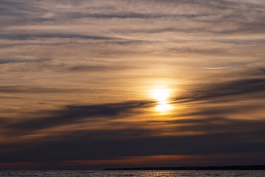 Golden sunset casts warm light across a calm sea as layered clouds stretch over the distant horizon, soft reflections shimmer on the water, creating a serene, tranquil coastal mood