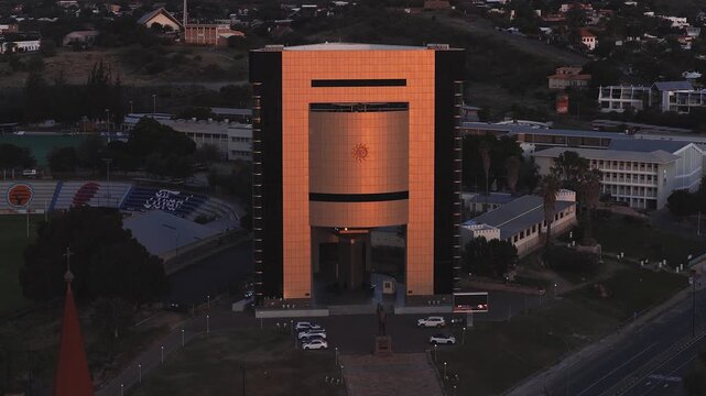 Drone captures the gold clad Independence Memorial Museum in Windhoek, with sun emblem, statue on stepped plinth, church spire, and stadium seating in warm light.