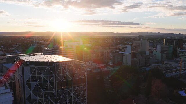 Aerial sunrise over Windhoek, Namibia. The drone pans as sun rays and lens flares light a geometric high rise, mid rise offices, tree lined parks, and distant low hills.