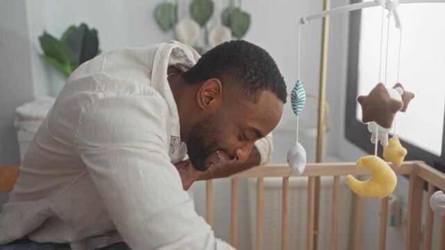 Young man smiling and leaning over wooden crib while playing with plush mobile in baby nursery; tenderness bonding.