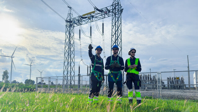 Three electrical engineers in helmets, safety harnesses and high-visibility workwear inspect a power substation beside wind turbines, supporting grid operation and renewable energy production.