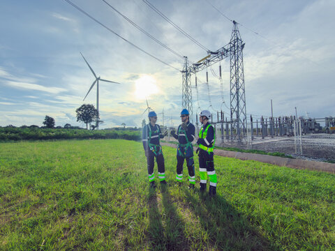 Three electrical engineers in helmets, harnesses and high-visibility workwear discuss a site inspection beside a power substation and wind turbines, supporting safe renewable electricity.