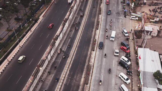 Drone aerial view of a busy city intersection featuring an underpass, multiple traffic lanes, cars, motorcycles, pedestrian crossings, and urban road infrastructure. The image represents transportatio