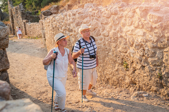 Senior couple tourists with hiking poles and hats exploring ancient stone ruins on summer vacation
