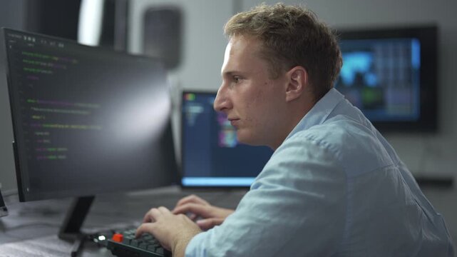 Male software engineer writing program code on multiple desktop screens in a modern office.