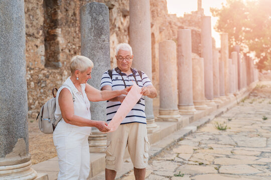 Senior couple tourists reading paper map at ancient Roman ruins with stone columns on summer vacation