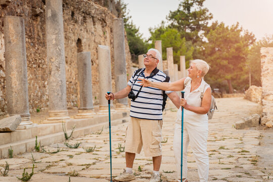 Senior couple tourists with hiking poles and hats exploring ancient stone ruins on summer vacation