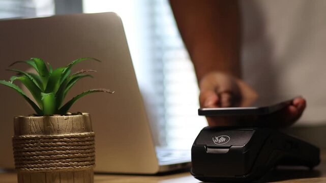 Customer using smartphone for contactless payment on a pos terminal at a wooden counter with a laptop in the background