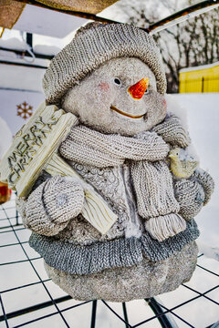 A snowman stands outside, holding a sign that says Merry Christmas. A festive Christmas tree is nearby, surrounded by snow