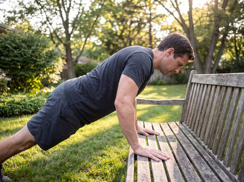 Fit young man doing push ups on a wooden park bench in sunlit backyard