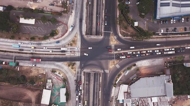 Drone aerial view of a busy city intersection featuring an underpass, multiple traffic lanes, cars, motorcycles, pedestrian crossings, and urban road infrastructure. The image represents transportatio